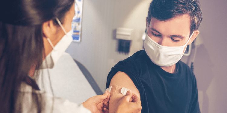 A healthcare worker applies a bandage to a man's upper arm after administering a vaccination. Both are wearing face masks.