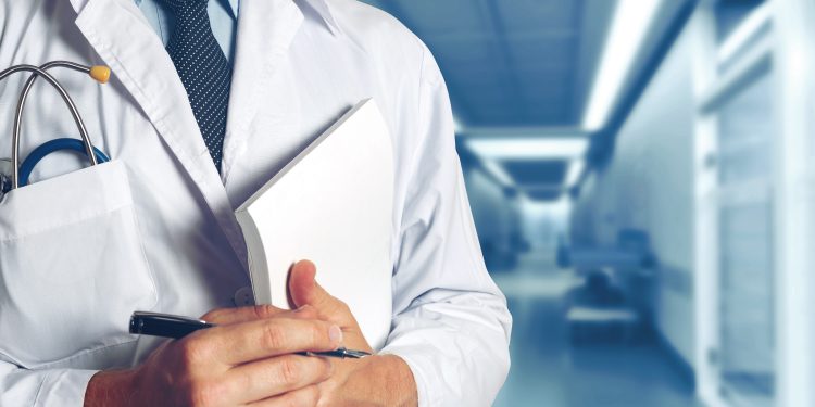 A doctor in a white coat holding a notepad and pen stands in a brightly lit hospital corridor.