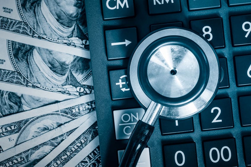 A stethoscope rests on a calculator next to several US hundred dollar bills, symbolizing healthcare costs or financial analysis in medicine.