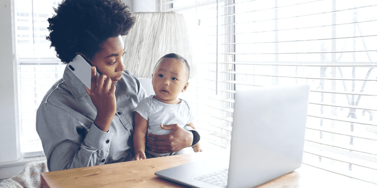 A person talks on the phone while holding a baby and sitting in front of an open laptop at a table.