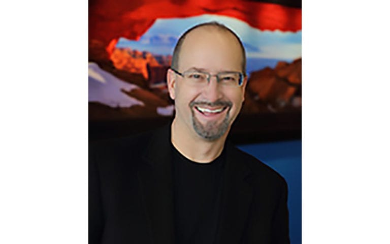 A man with glasses and a goatee smiles in front of a colorful, blurred landscape photograph.