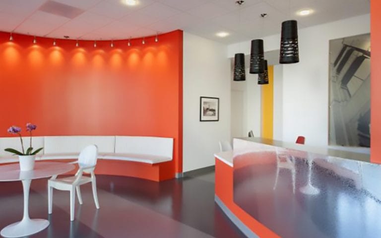 Modern office lobby with orange accent wall, white curved seating, a round table with a plant, and a reception desk with black pendant lights overhead.