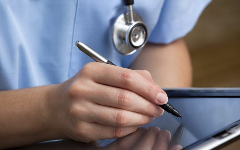 A person in medical scrubs with a stethoscope uses a stylus to write on a tablet.
