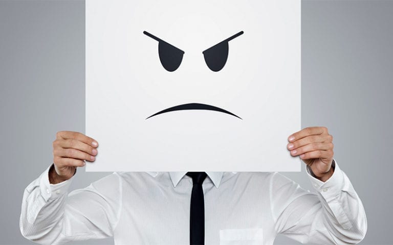 A person in a white shirt and tie holds a large sign with an angry face drawn on it, covering their head.
