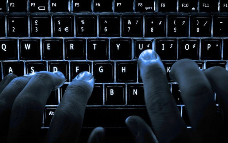 Close-up of hands typing on an illuminated QWERTY keyboard in low light.