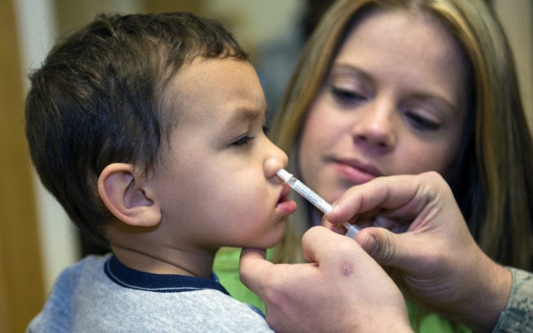 A young child receives a nasal spray while being held by an adult woman. Another person's hands are administering the spray.