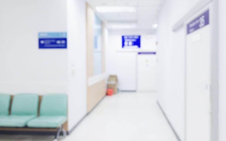 A brightly lit, modern hospital corridor with white walls, a row of light green chairs, and signs above doors; the image is slightly out of focus.
