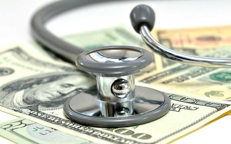 A stethoscope rests on top of several U.S. dollar bills, symbolizing the connection between healthcare and financial costs.