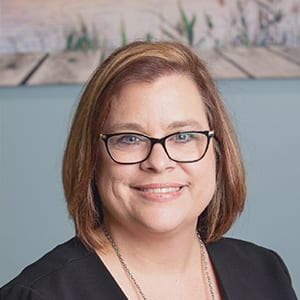 A woman with shoulder-length brown hair and glasses is smiling, wearing a black top and necklace, with a blurred background featuring a landscape photo.
