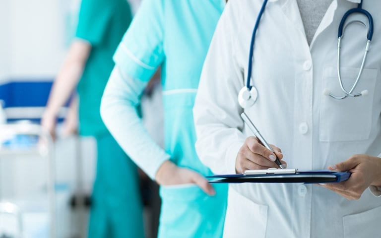 A doctor in a white coat writes on a clipboard, while a nurse in teal scrubs stands beside them in a hospital setting.