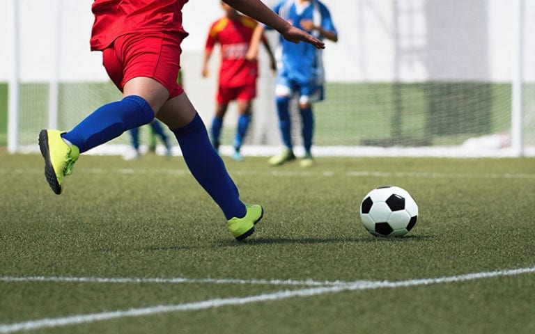 A soccer player in red and blue about to kick a black-and-white soccer ball on a field, with other players visible in the background.