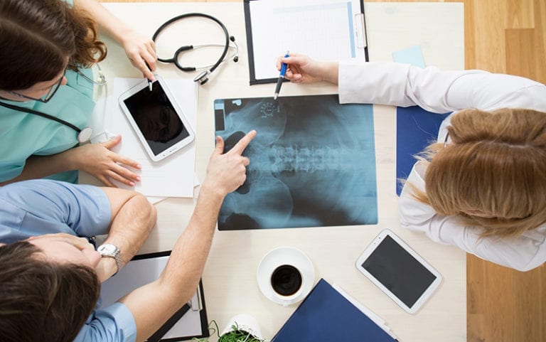 Three medical professionals review a spinal X-ray on a desk with tablets, a clipboard, a stethoscope, and a cup of coffee.