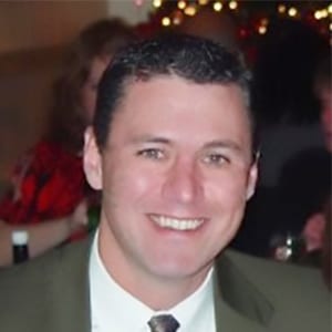 A man with short dark hair, wearing a suit and tie, smiles at the camera in a dimly lit indoor setting with blurred people and lights in the background.