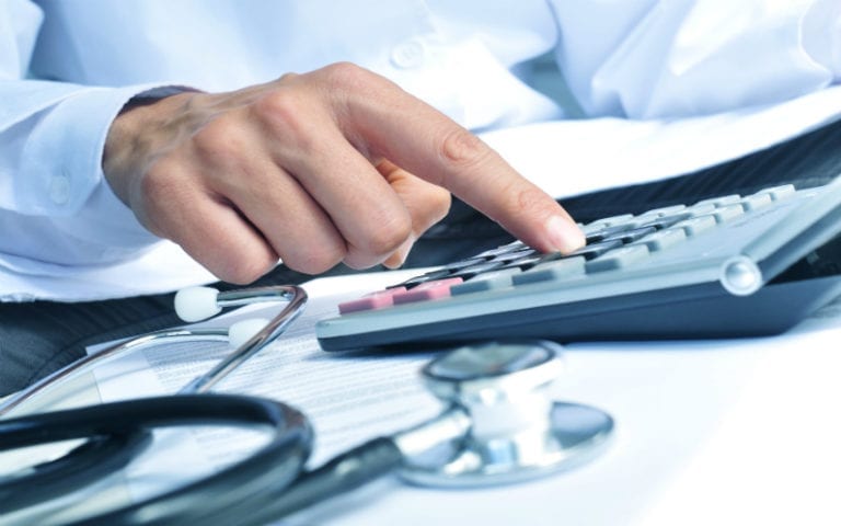 A person in a white coat uses a calculator, with a stethoscope and documents on the desk.