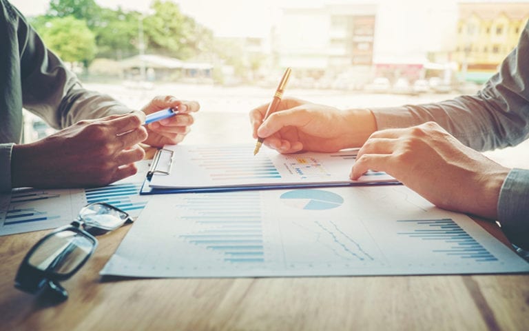 Two people review and discuss financial charts and graphs on printed documents at a desk, with pens and eyeglasses visible.