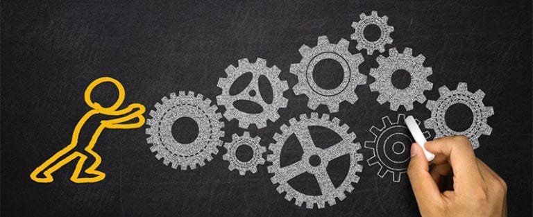 A chalk drawing of a person pushes a gear, which is part of a larger system of interconnected gears, on a blackboard. A hand draws one of the gears with white chalk.