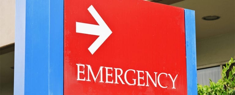 Red emergency sign with a white arrow pointing right, mounted outdoors with part of a building and some greenery in the background.