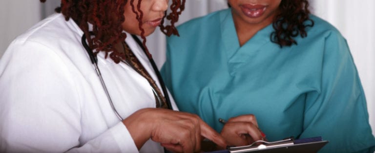 Two healthcare professionals review information on a clipboard; one is wearing a white coat with a stethoscope, and the other is in teal scrubs.