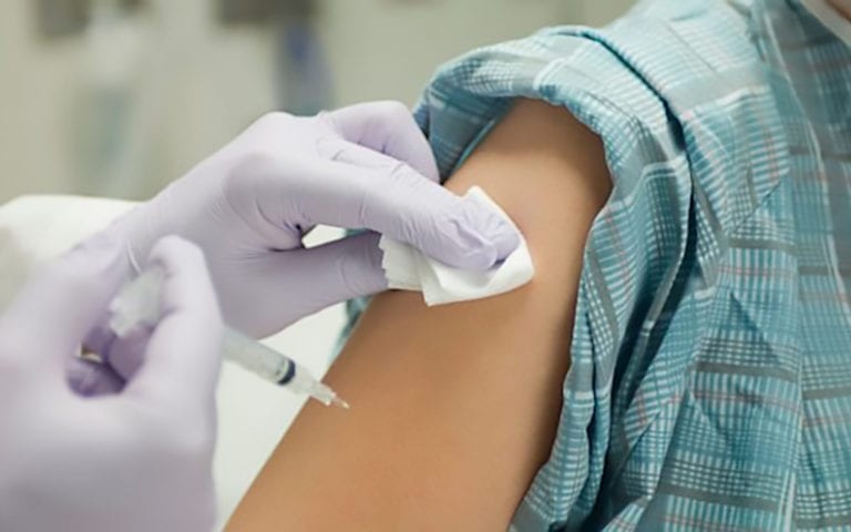 A person in a hospital gown receives an injection in the upper arm; a gloved hand holds a syringe while another cleans the skin with gauze.