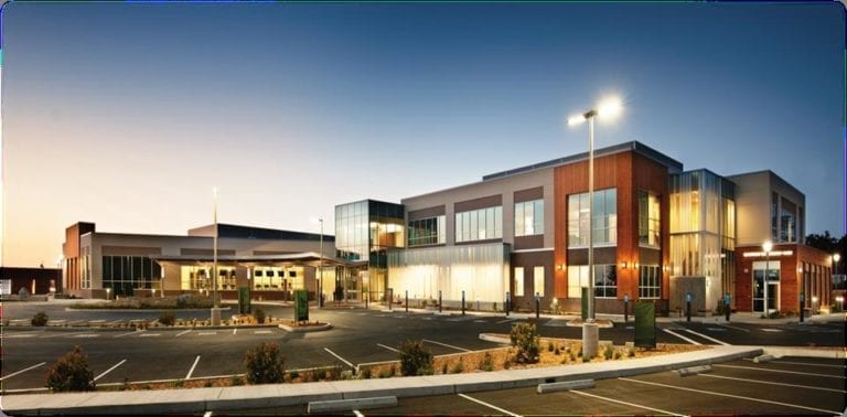 Modern hospital building with large glass windows and wood paneling, illuminated at dusk, with an empty parking lot in the foreground.