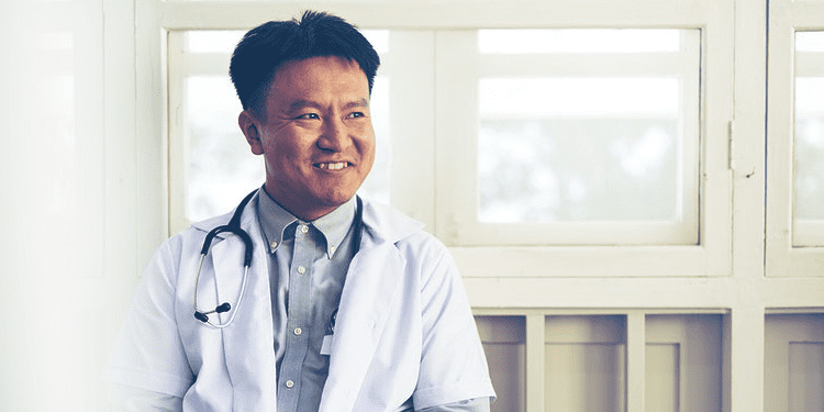 A man wearing a lab coat and stethoscope sits and smiles in front of large windows.