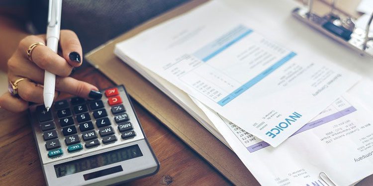 A person uses a calculator next to a clipboard holding invoices and documents on a wooden desk.