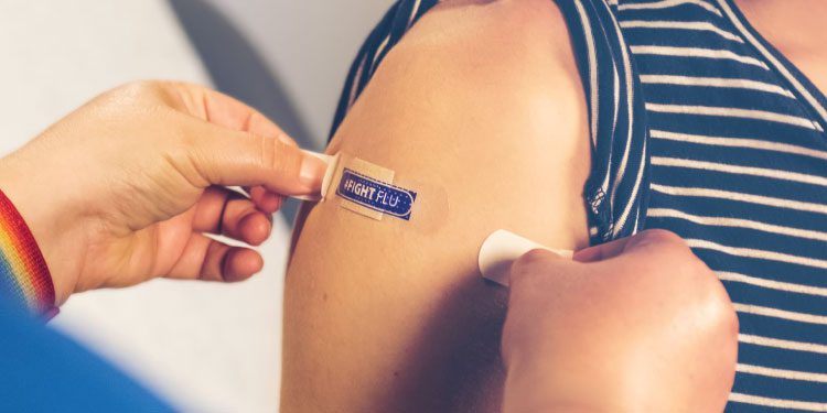 A person is applying a bandage labeled “I GOT MY FLU” to another person's upper arm after an injection.