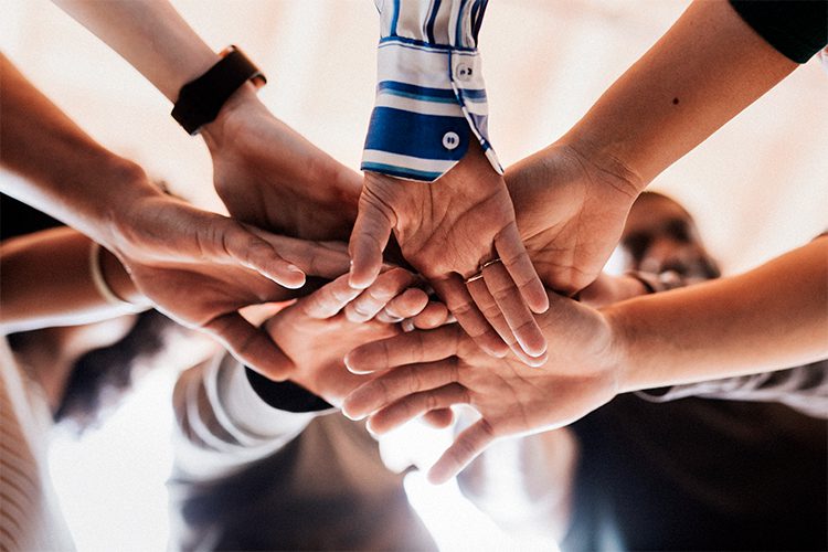 A group of people standing in a circle place their hands together in the center, showing unity and teamwork.