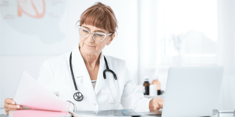 A doctor with a stethoscope reviews documents at a desk while looking at a laptop in a bright office.