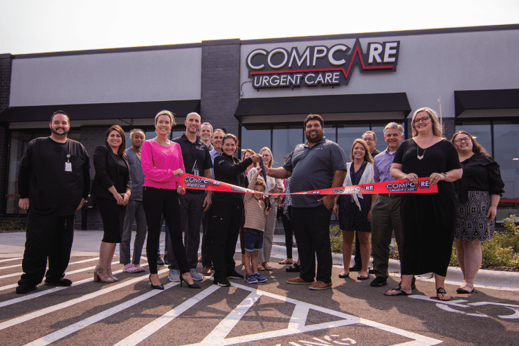 A group of people stands outside Compcare Urgent Care, smiling as two individuals cut a red ribbon at a grand opening event.