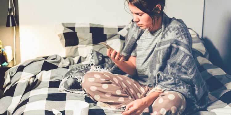 Woman sitting on a bed with a checkered blanket, wearing pajamas and a shawl, looking at her phone with a thermometer and mug nearby.