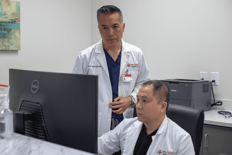 Two doctors in white coats, one standing and one seated, look at a computer monitor in a medical office setting.