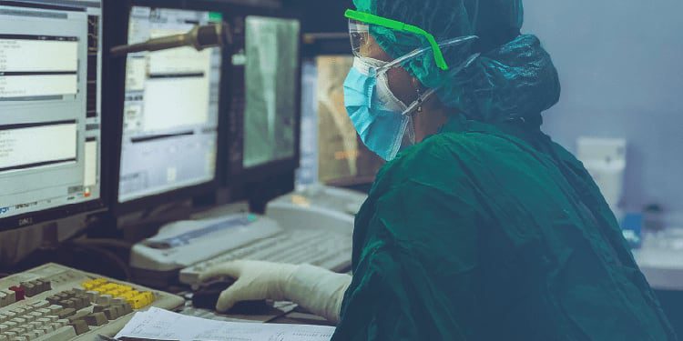 A medical professional in protective gear works at a computer station with multiple monitors, reviewing documents and data.