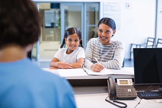 A woman and a young girl are at a reception desk, speaking with a staff member. The woman is filling out paperwork, and a phone is on the counter.