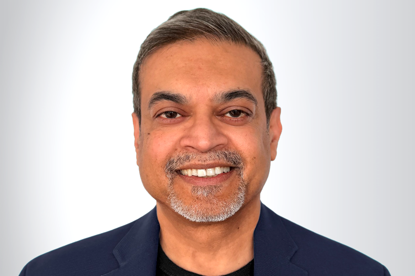 A man with short gray hair and a trimmed gray beard, wearing a dark blazer and a black shirt, smiles at the camera against a light gray background.