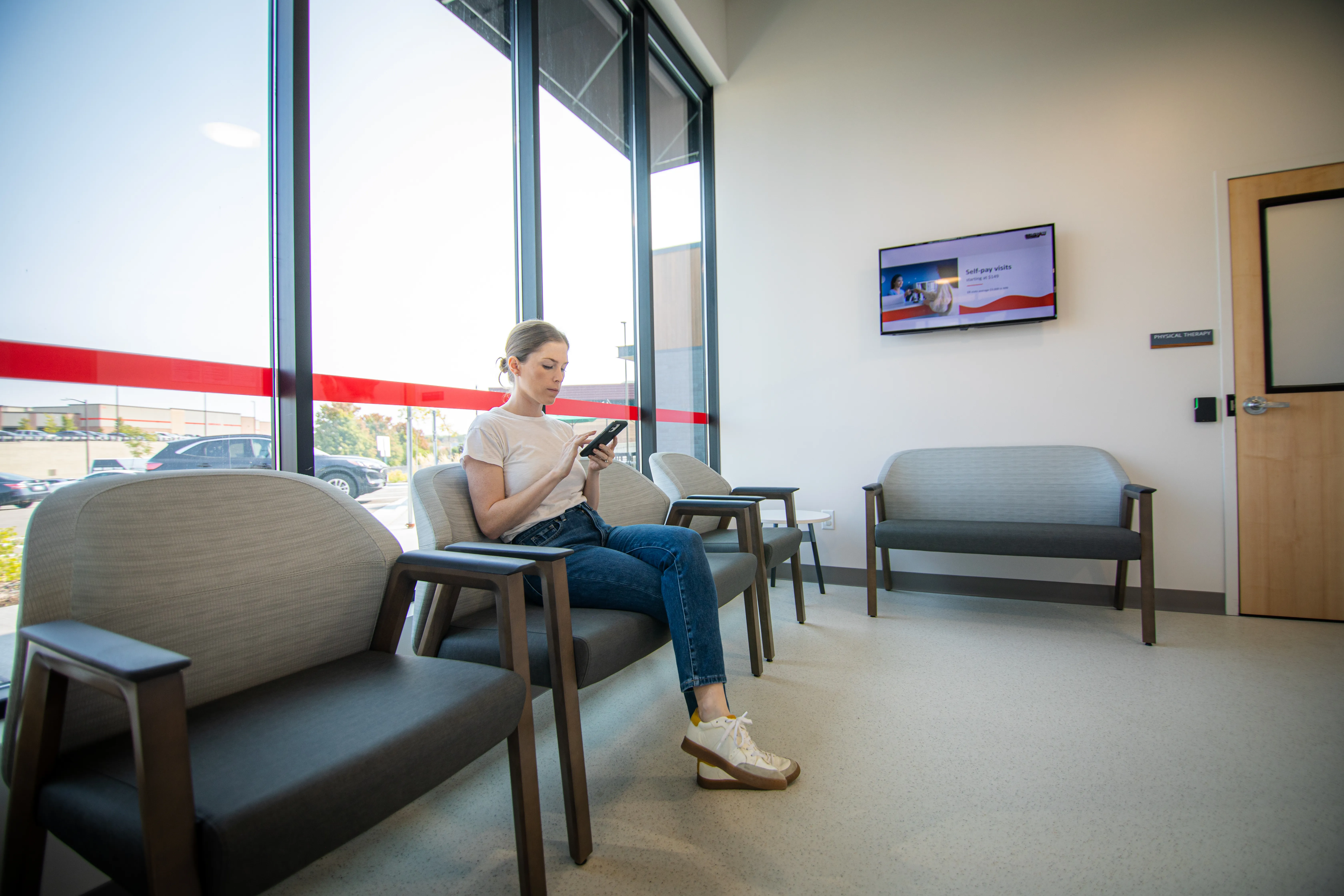 A woman sits alone in a modern waiting room, looking at her phone. There are empty chairs, a window with sunlight, and a wall-mounted TV in the background.