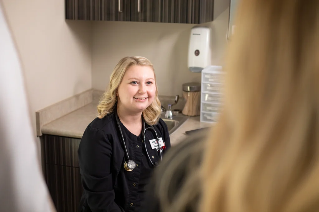 A healthcare professional in black scrubs with a stethoscope around her neck sits and smiles in a medical office, facing two people.