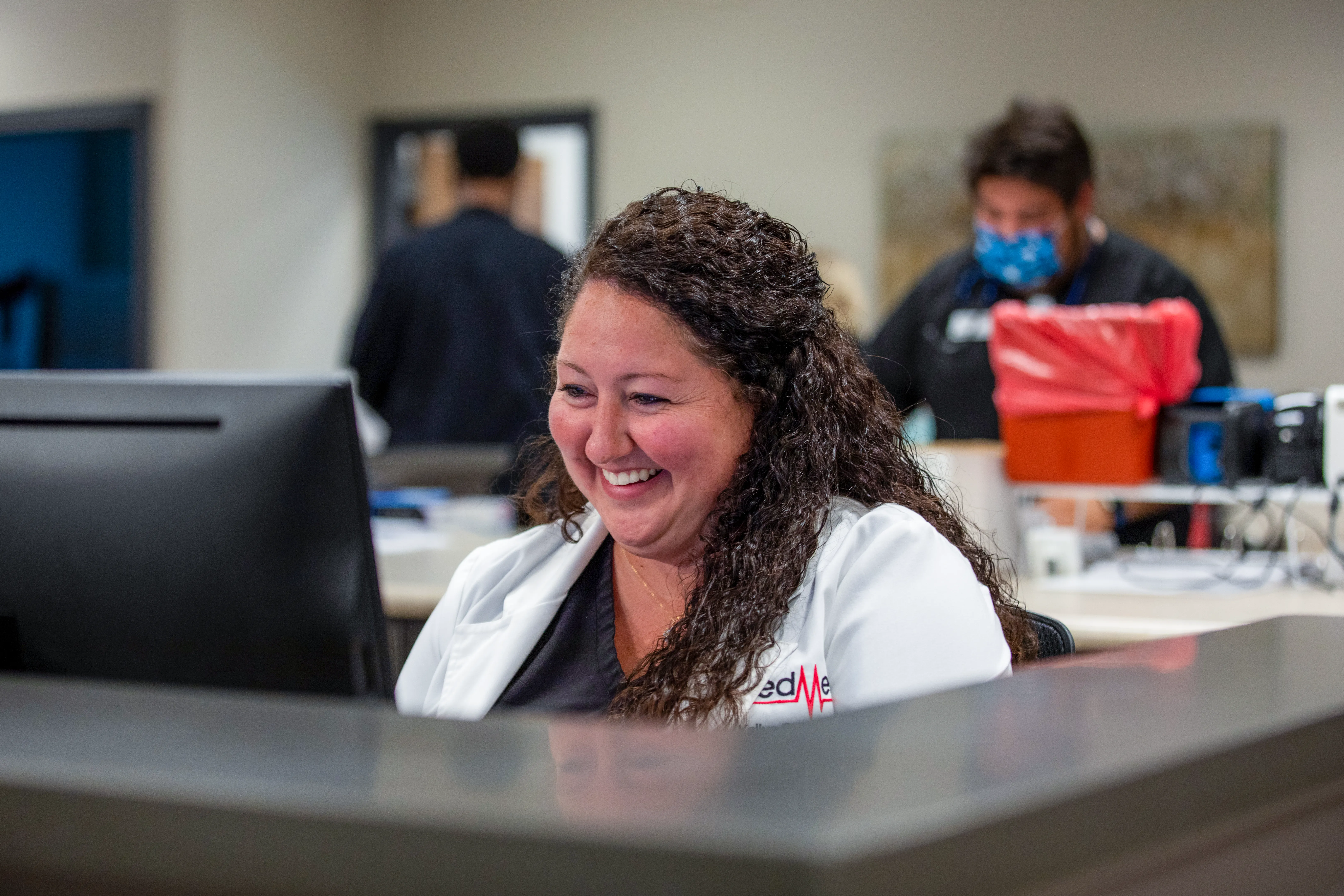 A woman in a white lab coat smiles while working at a computer in a medical office; two people work in the background.