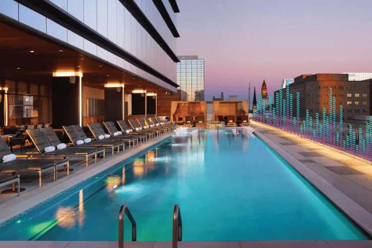 Outdoor rooftop pool lined with lounge chairs, city buildings in the background, and colored light patterns along the pool edge at dusk.