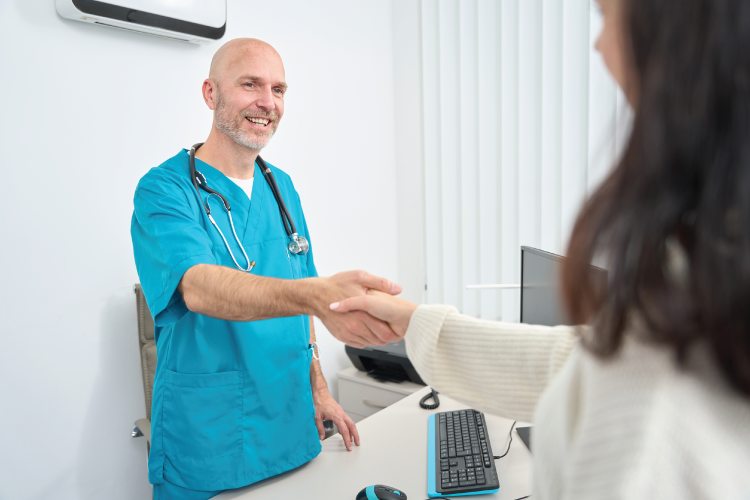 A doctor in blue scrubs smiles and shakes hands with a patient across a desk in a medical office.