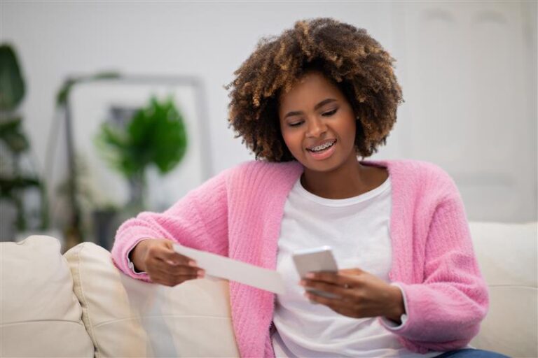 A woman with curly hair and braces sits on a couch, smiling while holding a piece of paper in one hand and a smartphone in the other.