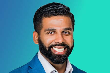A man with dark hair and a beard, wearing a blue suit jacket and white shirt, smiles in front of a blue to teal gradient background, reflecting the calm professionalism often valued in urgent care settings guided by the peak end rule.