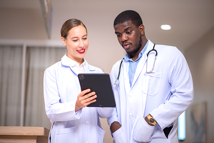 Two doctors in white coats stand together, looking at a tablet screen and discussing its contents in a medical office setting.
