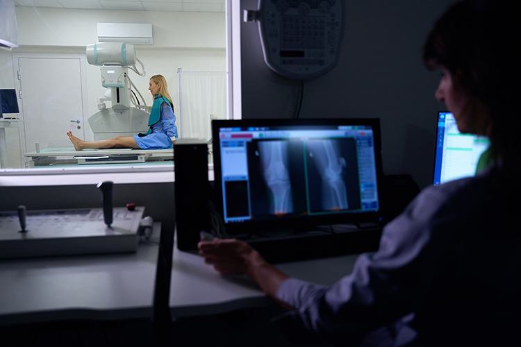 A patient sits for an X-ray while a technician analyzes knee and leg X-ray images on a computer in a control room.