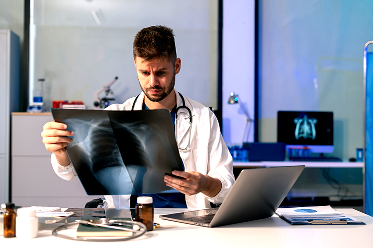 A doctor examines a chest X-ray in a medical office, with a stethoscope around his neck and medical equipment on the desk.