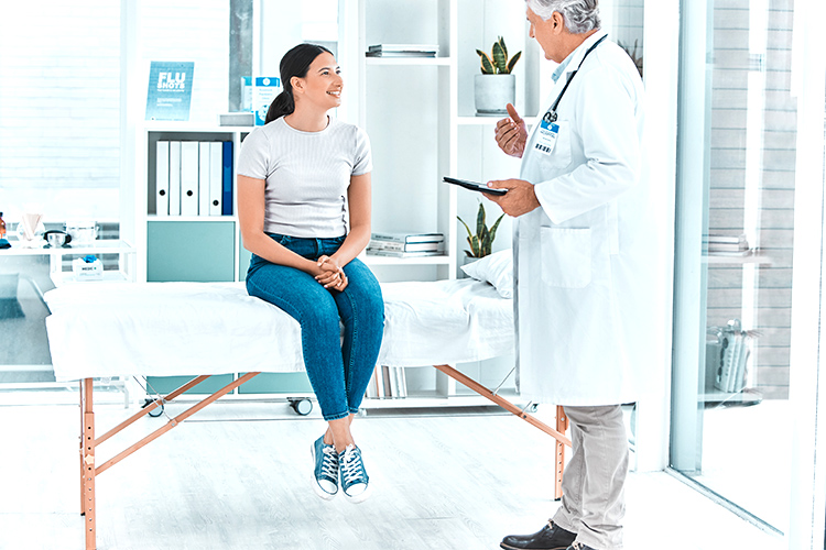 A woman sits on an exam table in a doctor's office, smiling and talking with a doctor who is holding a tablet.
