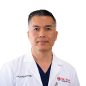 A man in a white medical coat with short dark hair stands against a plain white background.