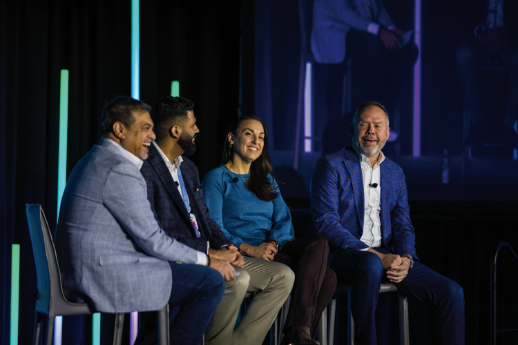 Four people sit on stage in chairs, engaged in a panel discussion about urgent care connect, with colorful vertical lights and a projected screen in the background.