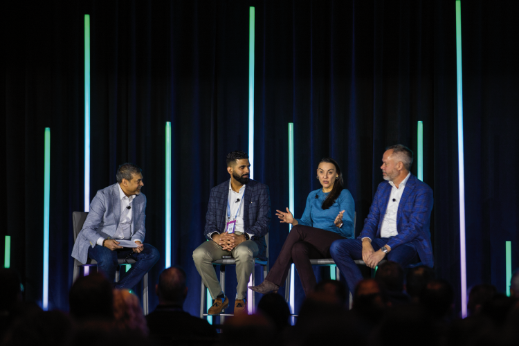 Four people sit on stage having a panel discussion in front of an audience, with vertical light bars in the background—exploring topics like Urgent Care Connect and its impact on healthcare access.