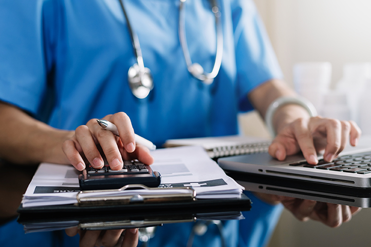 A person in medical scrubs uses a calculator and laptop at a desk, with paperwork and a stethoscope visible.